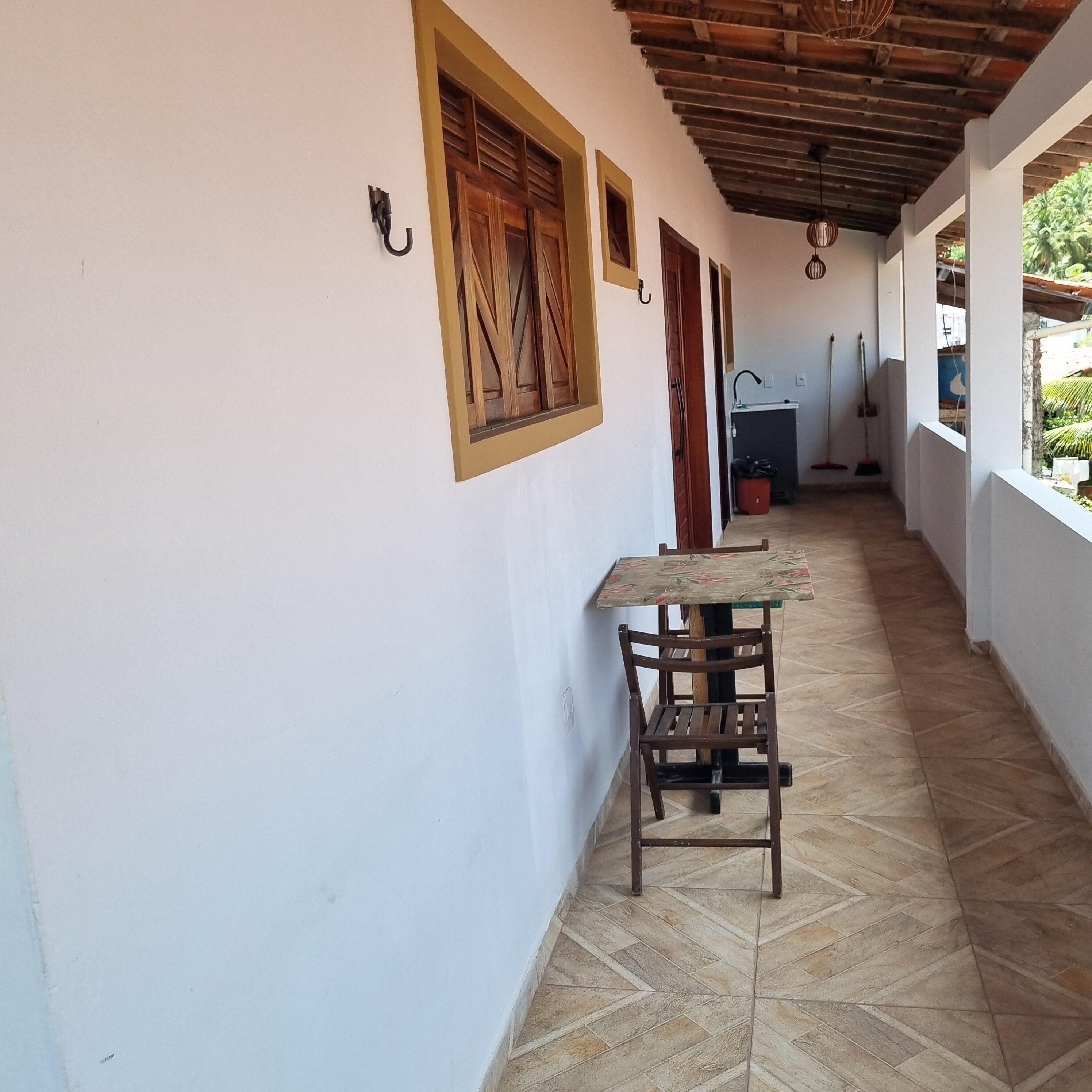 Um corredor com piso de azulejos, uma mesa de madeira com uma cadeira e uma janela com persiana de madeira.