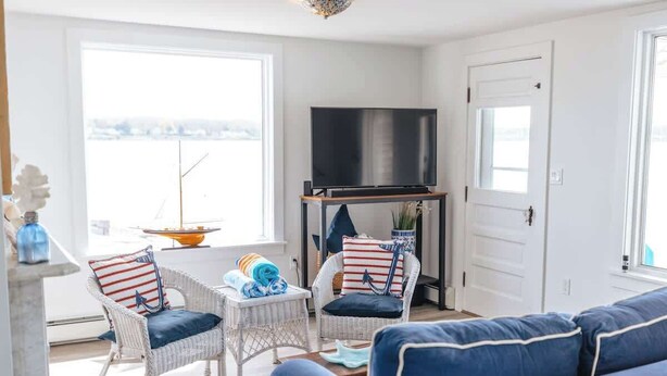 Een woonkamer met een bank, twee rieten fauteuils, een tv op een standaard en uitzicht door een raam.