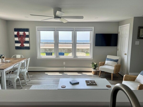 A modern living room with a dining area, large windows with a view of the beach, and a ceiling fan.