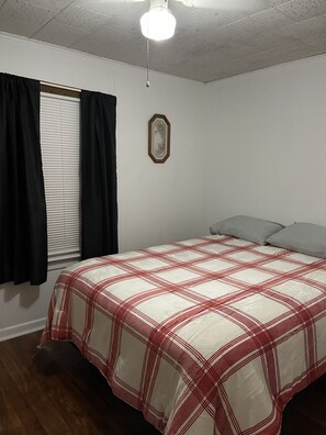 Une chambre à coucher avec un lit, une fenêtre avec des rideaux noirs, un ventilateur de plafond et un cadre photo fixé au mur.