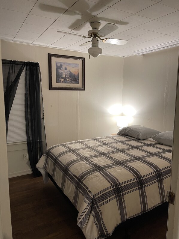 Une chambre à coucher avec un lit, un ventilateur de plafond, un tableau encadré au mur et une fenêtre avec des rideaux.