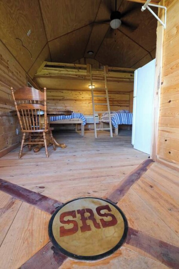 Interior de una cabaña con suelo de madera, literas, una mecedora, una escalera y una puerta con un cartel circular.