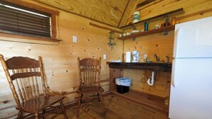 A wooden cabin interior with a dining area featuring two wooden chairs, a kitchen with a refrigerator, and a window with blinds. - Barn 2 @ Yellow Rose Canyon (Mount Enterprise)