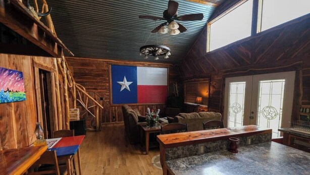 Ein rustikales Interieur mit Holzwänden, einer Texas-Flagge, einer Holztreppe, einer Bar und einem Sitzbereich mit Kamin.