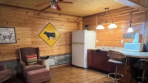 A rustic cabin interior with wooden walls, a ceiling fan, a kitchen area with a refrigerator and bar stools, and a yellow warning sign. - The Hideout @ Yellow Rose Canyon (Rusk County)