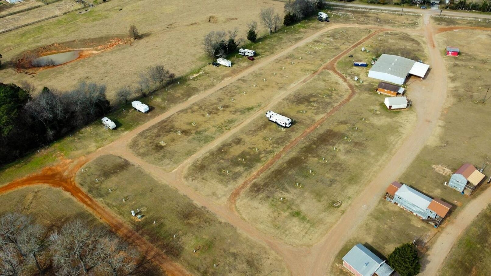 Vue aérienne d’une propriété rurale comprenant plusieurs parcelles rectangulaires, dont certaines sont occupées par des bâtiments et d’autres par des champs ouverts. On y aperçoit plusieurs remorques blanches et quelques petites constructions.