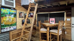A rustic cabin interior with a wooden table and chairs, a ladder leading to a loft, and a poster on the wall advertising a family reunion.