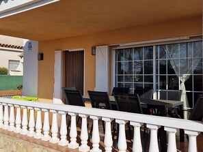 En udendørs terrasse med hvid gelænder, et bord med stole og et vindue med gardiner.