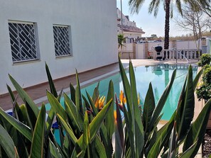 A swimming pool with surrounding plants, a white building with barred windows, and a clear sky.