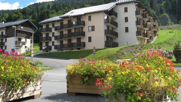 Un edificio de varios pisos con balcones, rodeado de macizos de flores y con montañas al fondo.