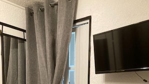 Una habitación con cortina gris, televisor montado, un cuadro en la pared y una pequeña mesa de madera.
