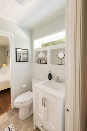 A bathroom with a white toilet, sink, and cabinet, a mirror, and a framed picture on the wall. - Cute n' Comfy Capers (Columbia)