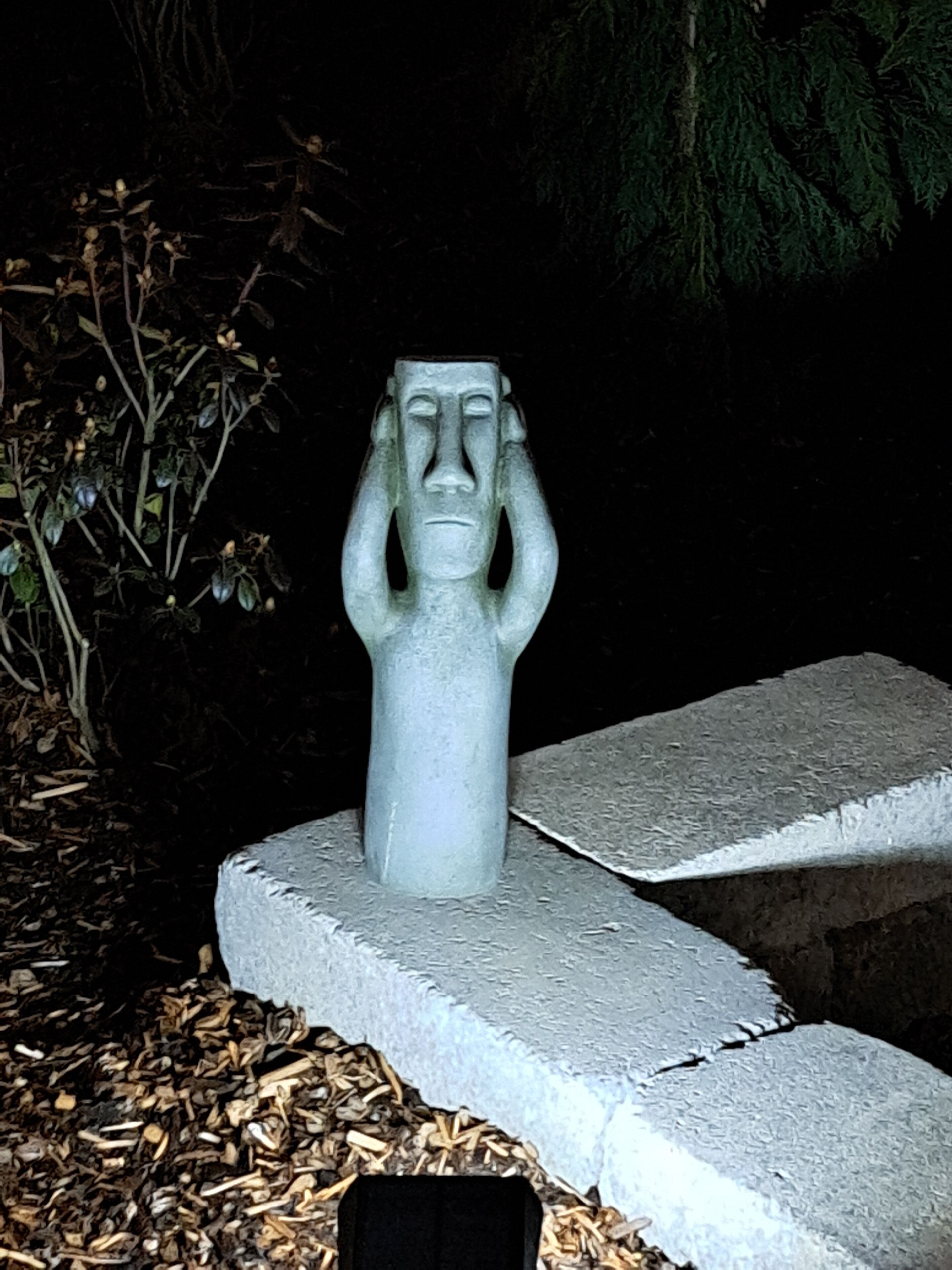 Una estatua de piedra con rostro, sobre un pedestal de concreto rodeado de mantillo y plantas.