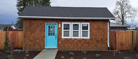 A small wooden house with a blue door, a front porch, and a small garden area.