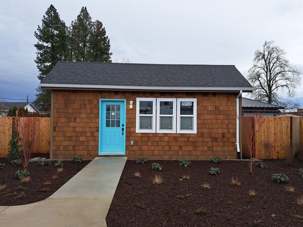A small wooden house with a blue door, a front porch, and a small garden area.