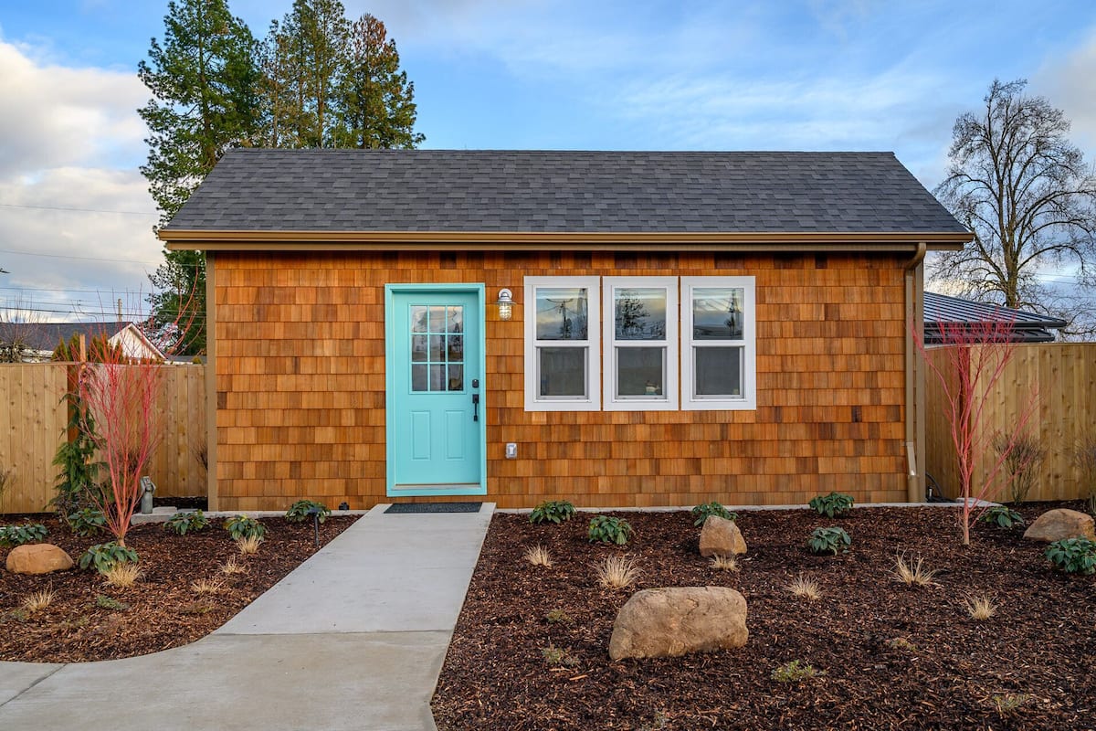 A small wooden house with a blue door, a front porch, and a landscaped garden with mulch and rocks.