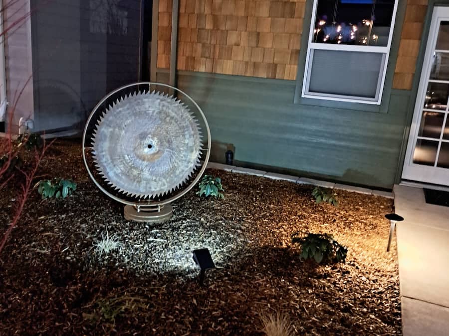 A circular saw blade in a garden area with mulch, a shed, and a house with a window and a door.