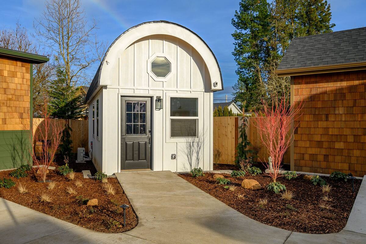 A small, white, semi-circular structure with a door and windows, surrounded by a wooden fence and a landscaped garden.