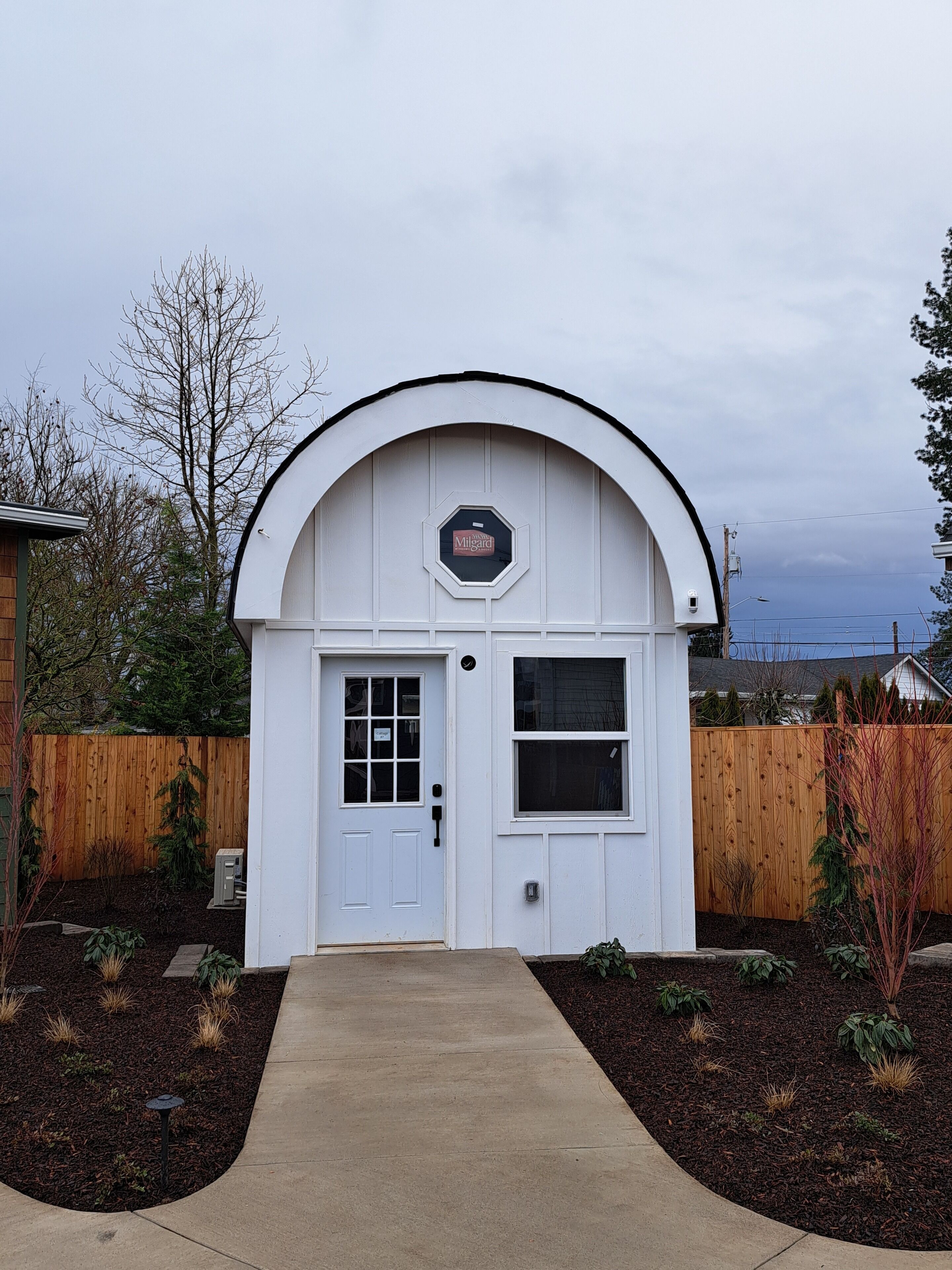 A small, white, arched building with a door and windows, surrounded by a wooden fence and a garden.