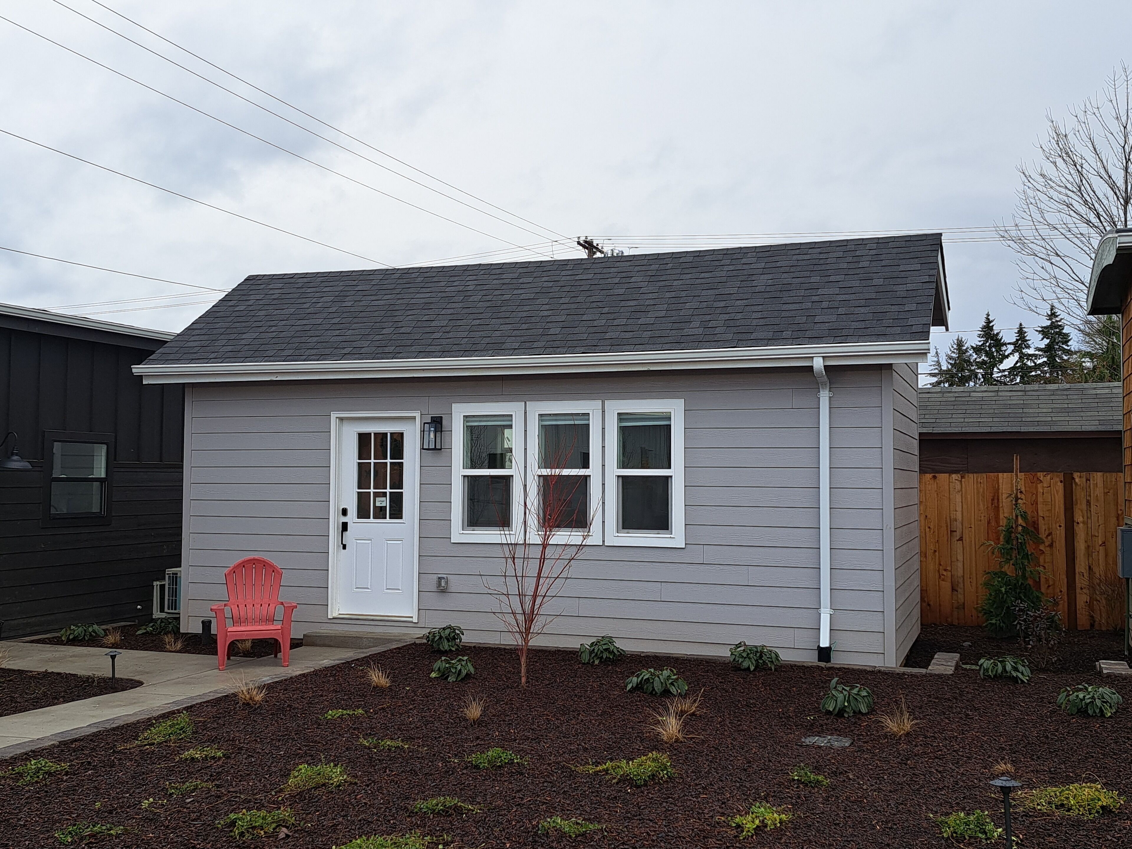 Une maison de plain-pied avec un revêtement extérieur gris, une porte d’entrée blanche et deux fenêtres. On peut voir une chaise Adirondack rouge à l’extérieur, ainsi qu’un petit arbre dans le jardin.