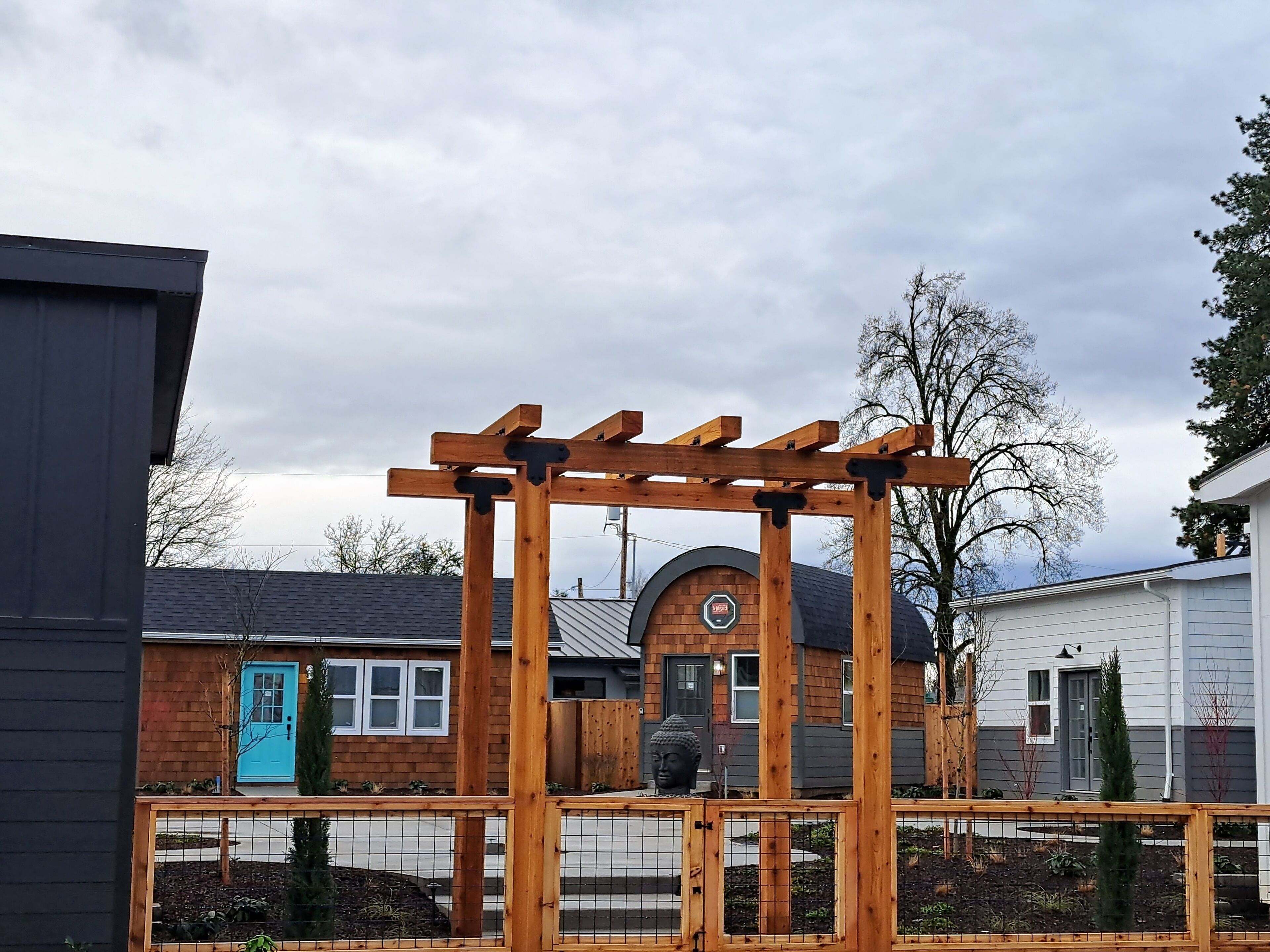 A wooden pergola structure in a residential area with a brick house, a blue door, and a circular building in the background.