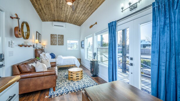 Un salón acogedor con un sofá de cuero marrón, una mesa de centro de madera y una alfombra estampada. Hay una chimenea, un ventilador de techo y un ventanal con cortinas azules.
