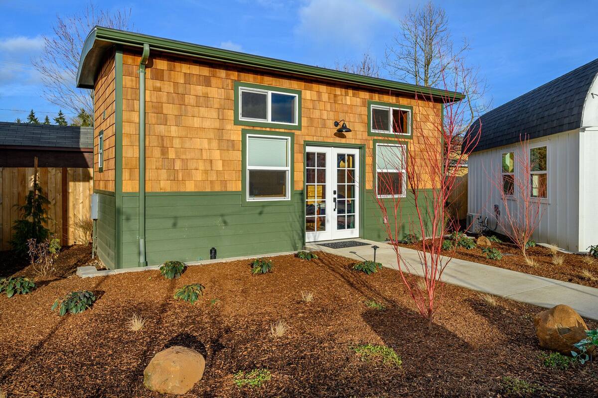 A two-story house with a green and brown exterior, a front door with glass panels, and a small front yard with mulch and plants.