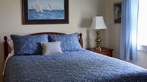 Una cama bien hecha con una colcha azul estampada, un cuadro enmarcado con barcos a vela en la pared y una lámpara de noche.