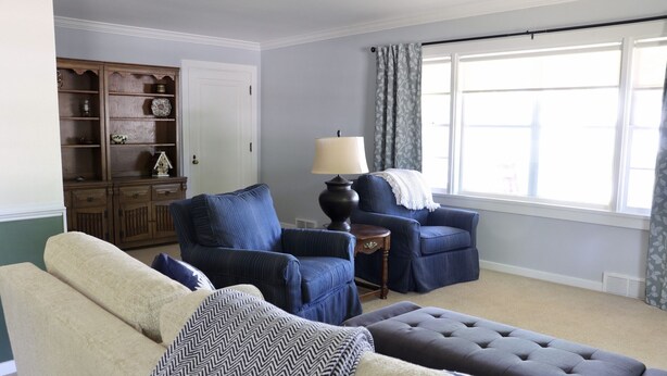 Una sala de estar con un sofá color crema, un sillón azul, un reposapiés oscuro, un mueble de madera y una lámpara.