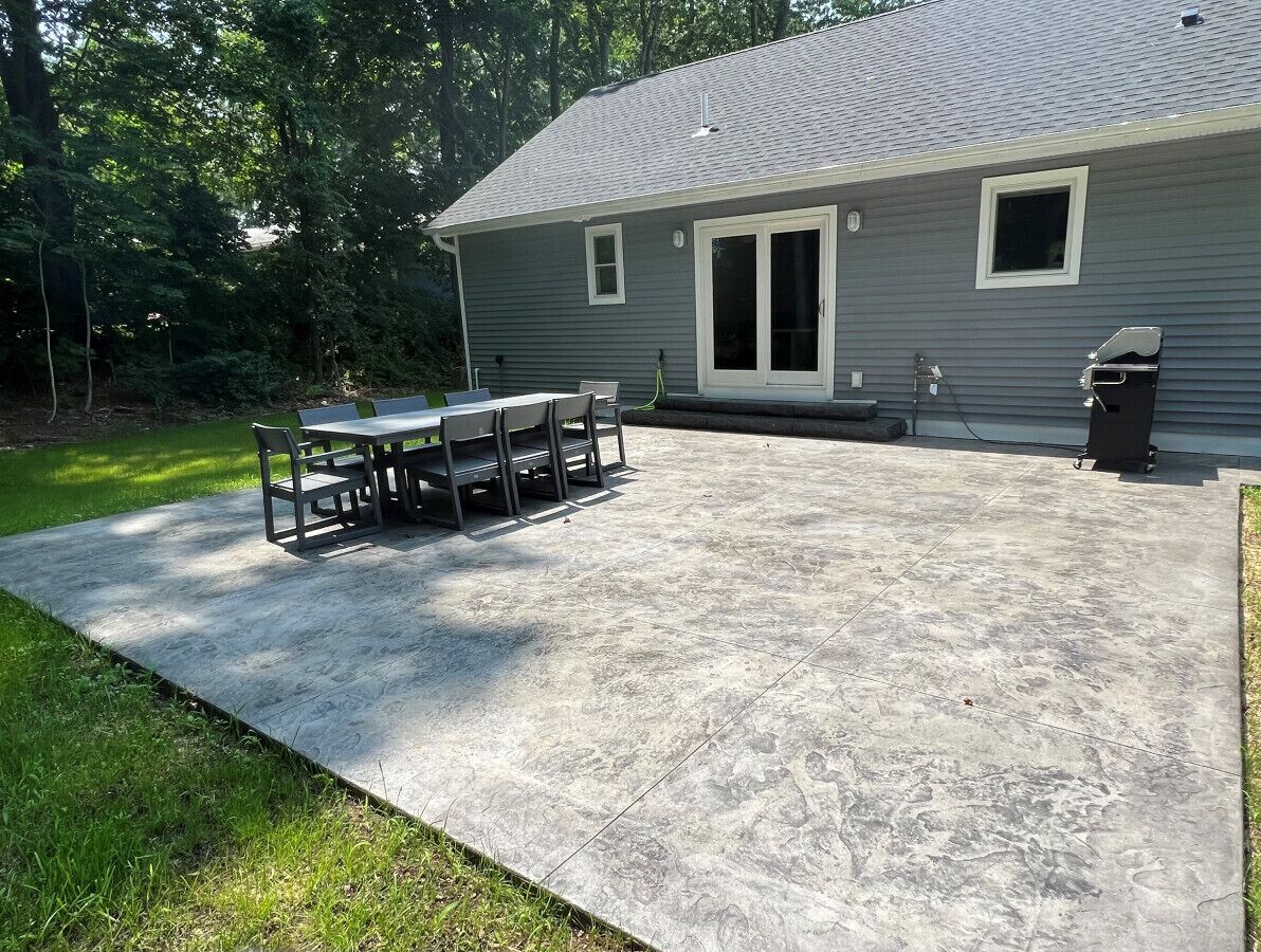 A concrete patio with a table and chairs, a covered area, and a house with a gray exterior and a garage door.