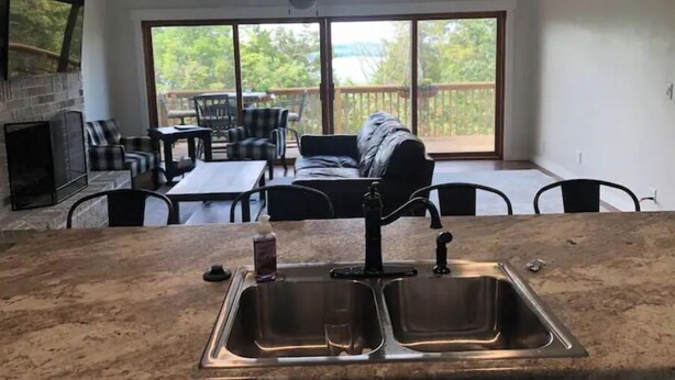 Een keuken met een dubbele spoelbak, een eethoek met een tafel en stoelen, een open haard en een balkon met uitzicht op bomen.