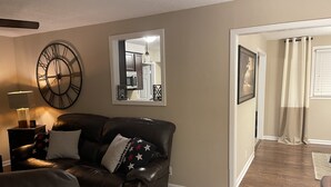 A living room with a dark brown leather sofa, a side table with a lamp, a large wall clock, and a mirror reflecting a kitchen area. - Home away from home 37043 (Clarksville)