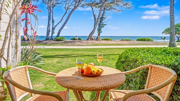 A wicker table with a glass of wine and fruit, overlooking a beach with trees and a clear sky. - View Beachfront, Split AC, Pool Hot Tub, 1st Floor (Kapaa)