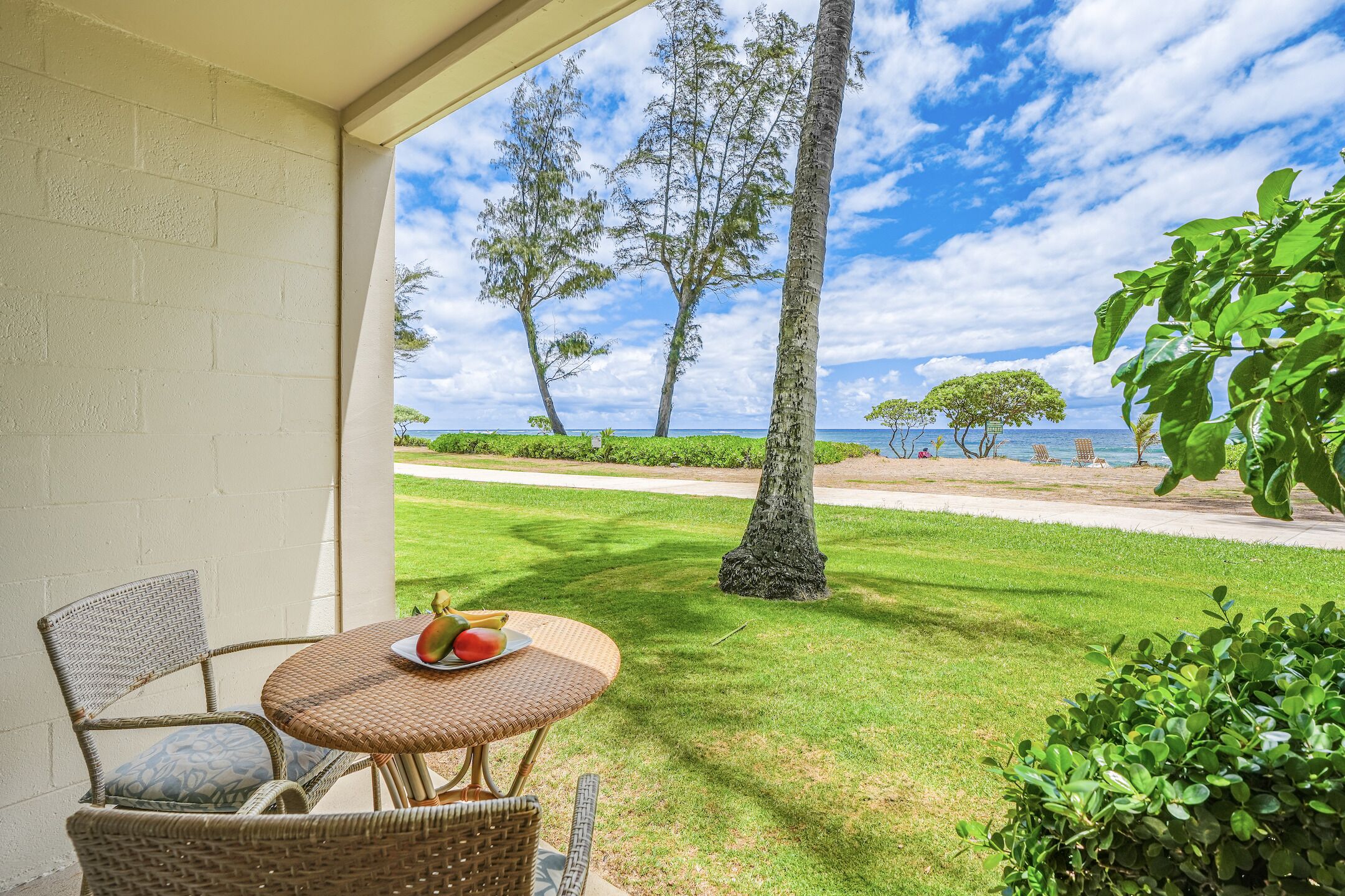 A patio with wicker furniture and a table with fruit, overlooking a grassy area with trees and a view of the sea.