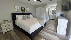 Un dormitorio con cama, mesitas de noche, ventilador de techo, cómoda, televisor y un baño visible al fondo.