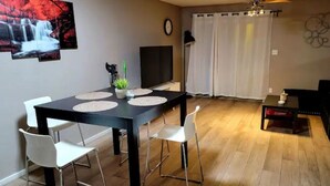 Ein Essbereich mit einem schwarzen Tisch, weißen Stühlen und einer Pflanze als Mittelpunkt. Ein Fernseher ist an der Wand befestigt, und eine Uhr ist sichtbar.