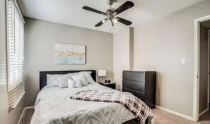 Un dormitorio con una cama, un ventilador de techo, una mesita de noche, un aparador y un cuadro en la pared.