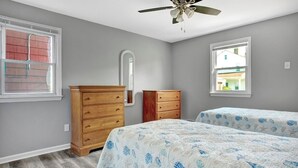 Un dormitorio con dos camas, cómodas de madera, un ventilador de techo y una ventana con vista a una casa.