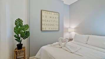 Una cama bien hecha con ropa blanca y un arreglo de toallas con forma de corazón, una planta en maceta y una cita motivacional en la pared.