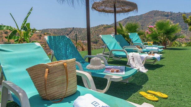 Une rangée de chaises longues turquoise, avec un sac en paille et un chapeau posés sur l’une d’elles, et une table sur laquelle se trouvent des boissons et un magazine.
