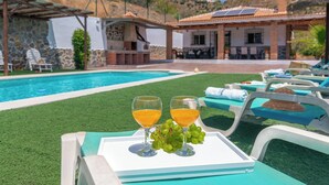 Une scène au bord de la piscine, avec deux verres de jus d’orange et une grappe de raisins sur un plateau, entourés de chaises longues et d’un mur de pierre.