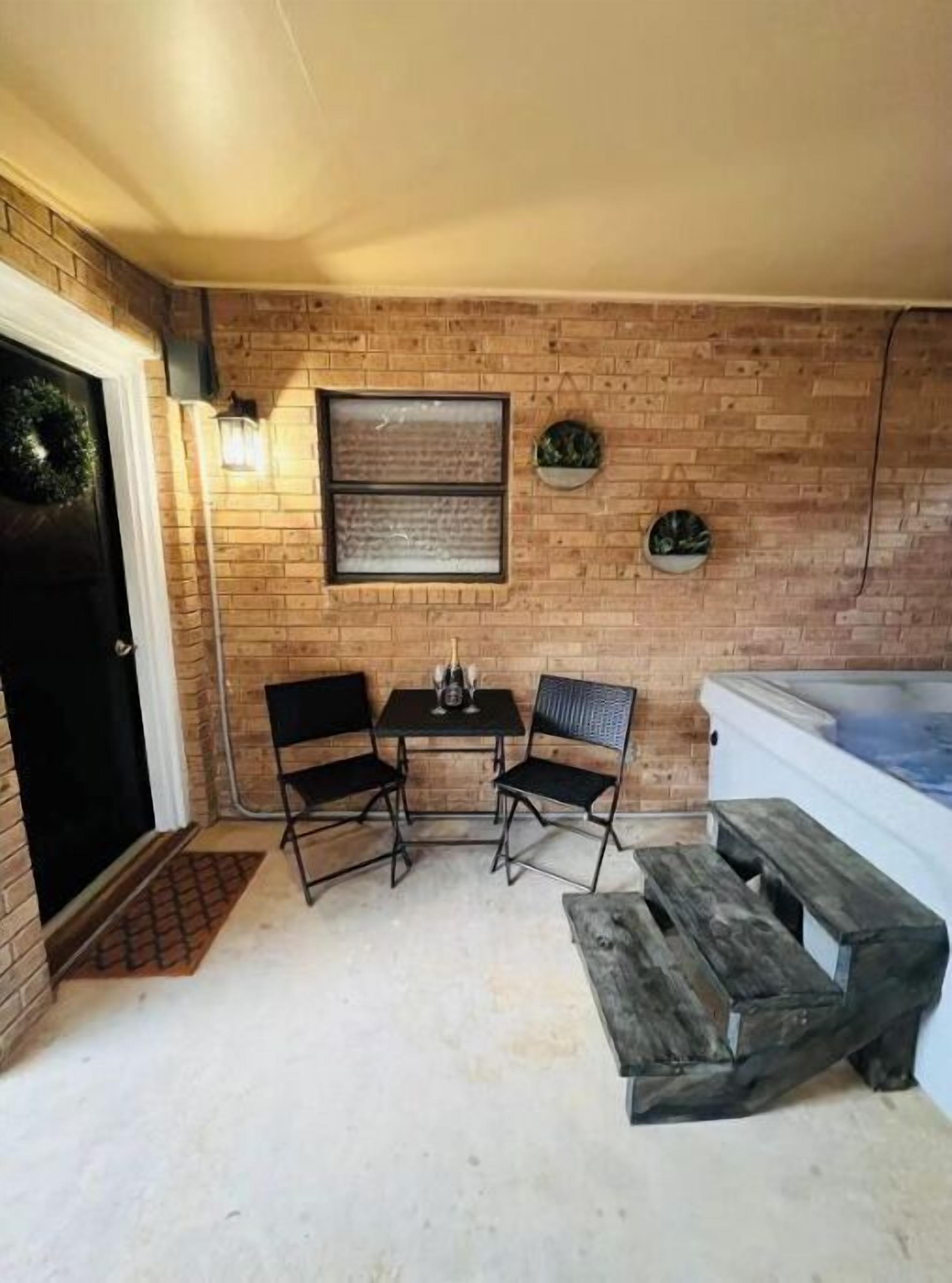 Pequeño patio amurallado con pared de ladrillo, puerta negra, una mesa pequeña con dos sillas y un jacuzzi.
