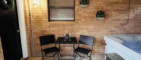 Pequeño patio amurallado con pared de ladrillo, puerta negra, una mesa pequeña con dos sillas y un jacuzzi.