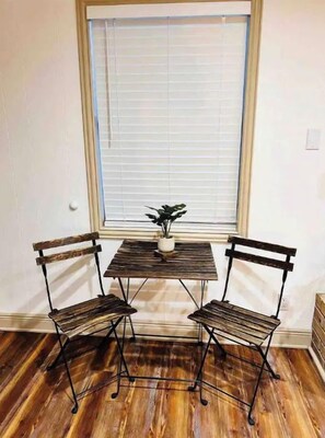 A corner room with a wooden floor, a small table with two chairs, and a window with blinds.