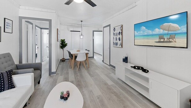 A modern living room with a ceiling fan, a dining area with a table and chairs, and a TV displaying a beach scene. - Shorehaven 2- Updated and Stylish 2 bedroom 14 St! (Ocean City)