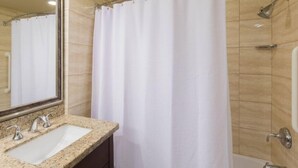 Ein Badezimmer mit Granitwaschtisch, einem Spiegel, einer Dusche mit weißem Duschvorhang und einer Haltestange.