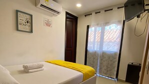 A hotel room with a bed, a television, an air conditioning unit, a door, and a window with curtains. - CL-7 Habitación con balcón en Getsemaní (Cartagena)