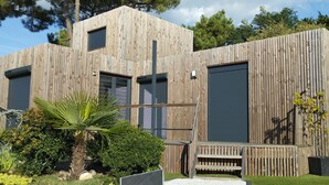 A modern wooden building with multiple garage doors, a small outdoor seating area, and a well-maintained garden with various plants. - Air-conditioned mini-house near the beaches, built in 2022 (Pornichet)