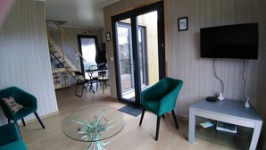 A living room with a glass table, two teal armchairs, a TV on a stand, and a staircase leading to an upper floor. - Air-conditioned mini-house near the beaches, built in 2022 (Pornichet)