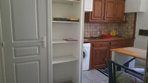 Kompakti keittiö, jossa on puiset kaapit, pesukone ja ruokailutila, jossa on yksi tuoli.